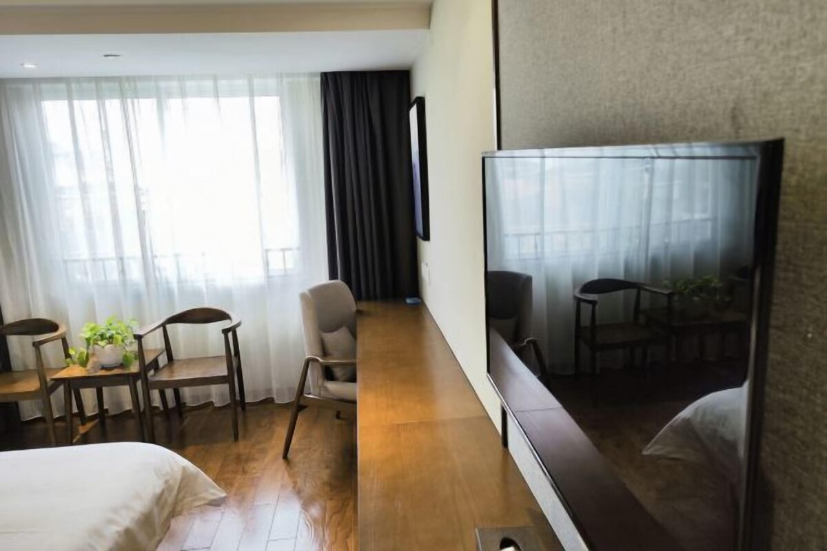 Habitación de hotel con cama, escritorio, silla, televisor y ventana grande con cortinas.