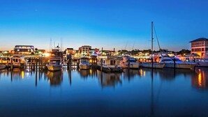 A marina at dusk with boats docked, waterfront buildings, and a clear sky.