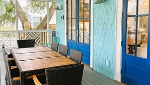 A covered outdoor dining area with a wooden table and wicker chairs, a blue door, and a view of the outdoors.