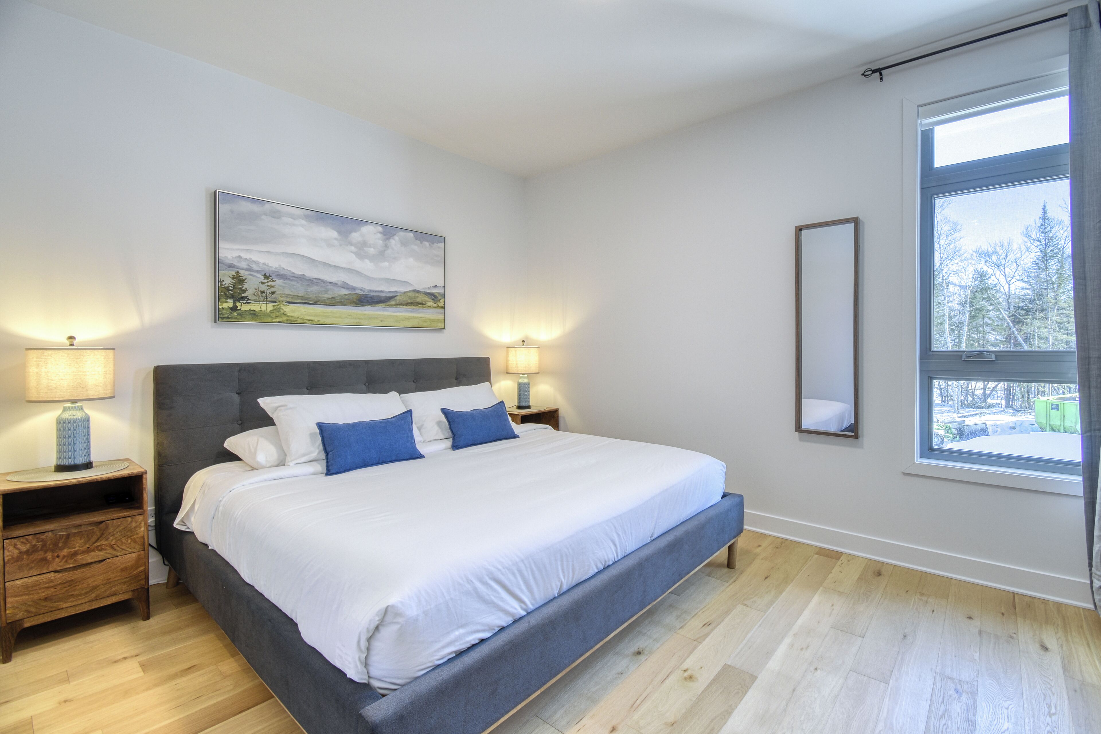 Une chambre à coucher avec un grand lit, deux tables de chevet, une commode, une fenêtre donnant sur un beau paysage et un tableau accroché au mur.