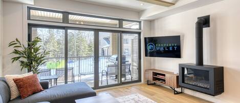 A modern living room with a grey sofa, a wooden coffee table, a flat-screen TV mounted on the wall, and a black wood-burning stove.
