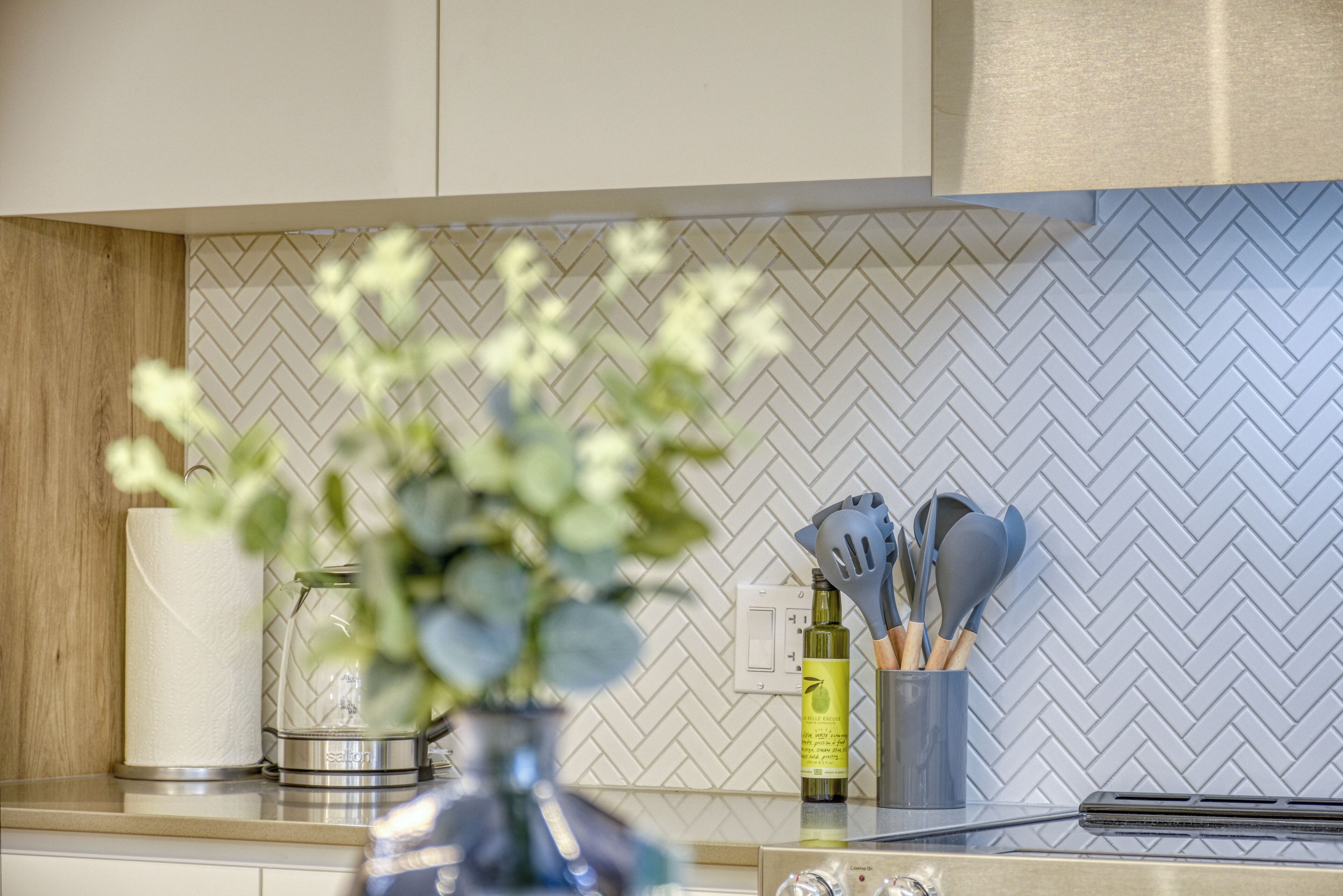 A kitchen with a herringbone tile backsplash, a vase with flowers, and kitchen utensils on the counter.
