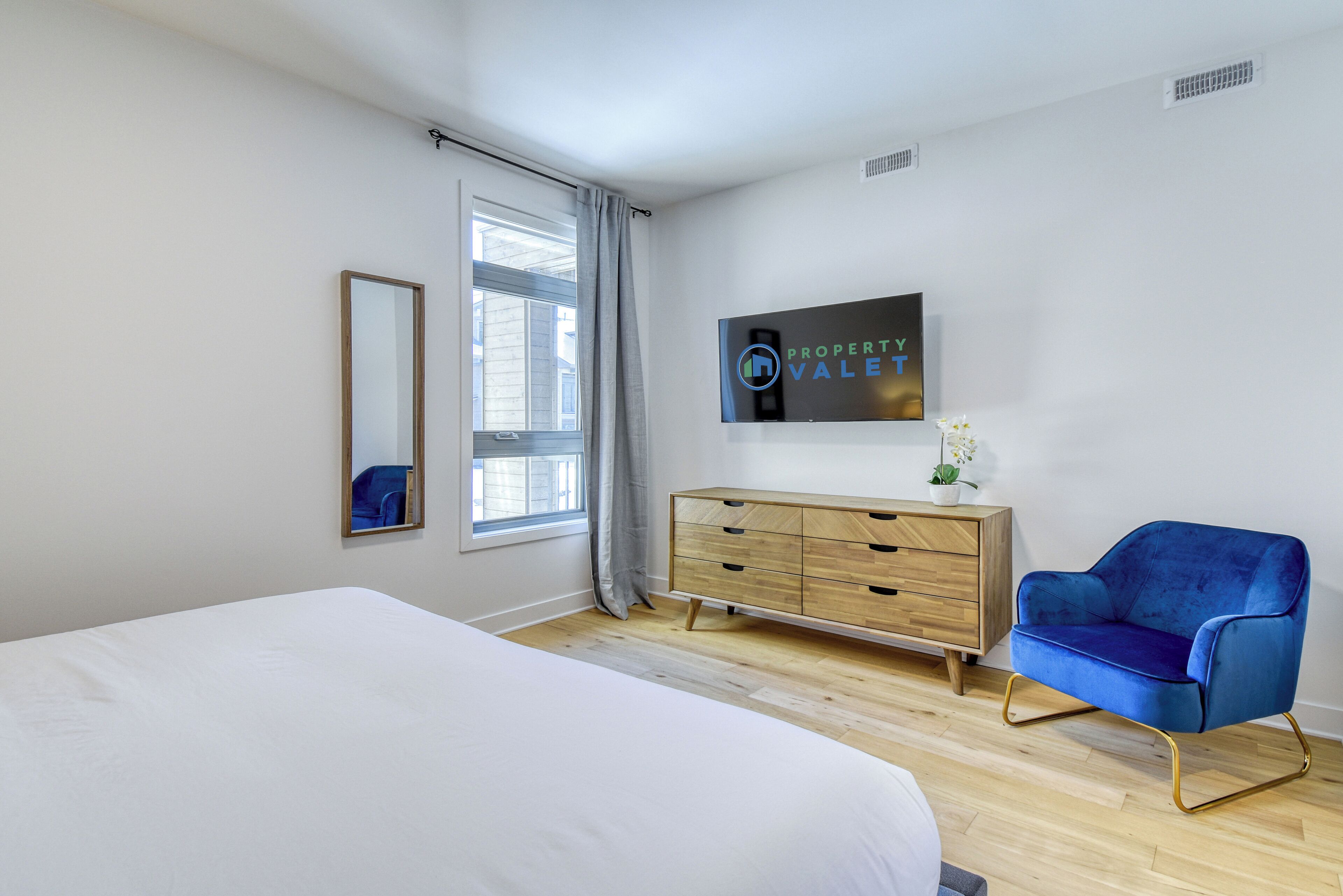 A modern bedroom with a large bed, a wooden dresser, a blue armchair, and a TV mounted on the wall.