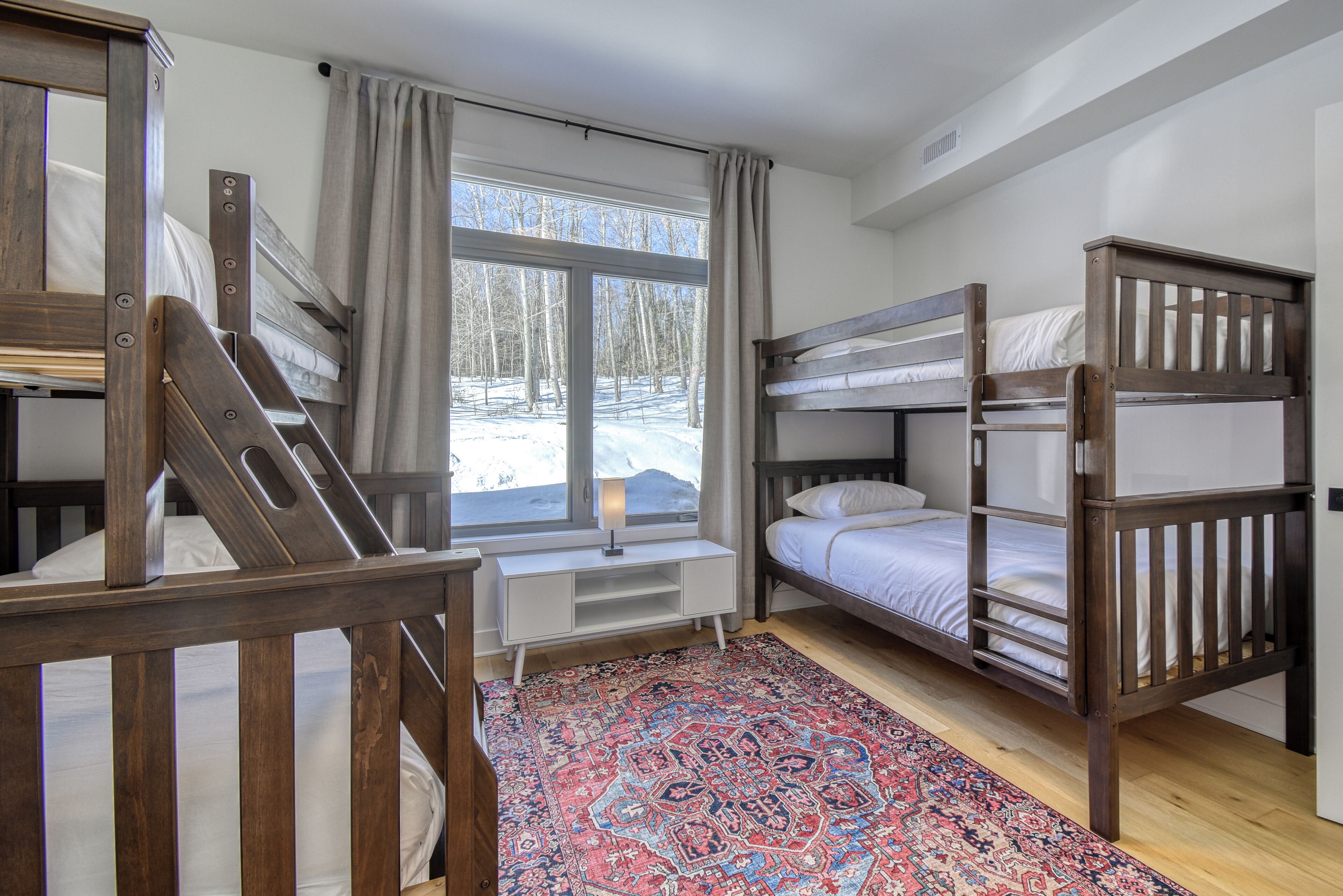 A room with bunk beds, a window with a view of a snowy landscape, a desk with a lamp, and a patterned rug.