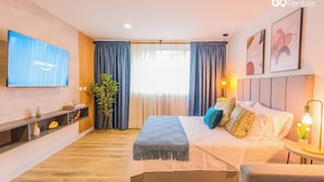 A modern hotel room with a large bed, a flat-screen TV, a bedside table with a lamp, and a large window with curtains.
