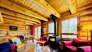 A cozy log cabin interior with a central fireplace, wooden walls, and large windows offering a view of the forest.