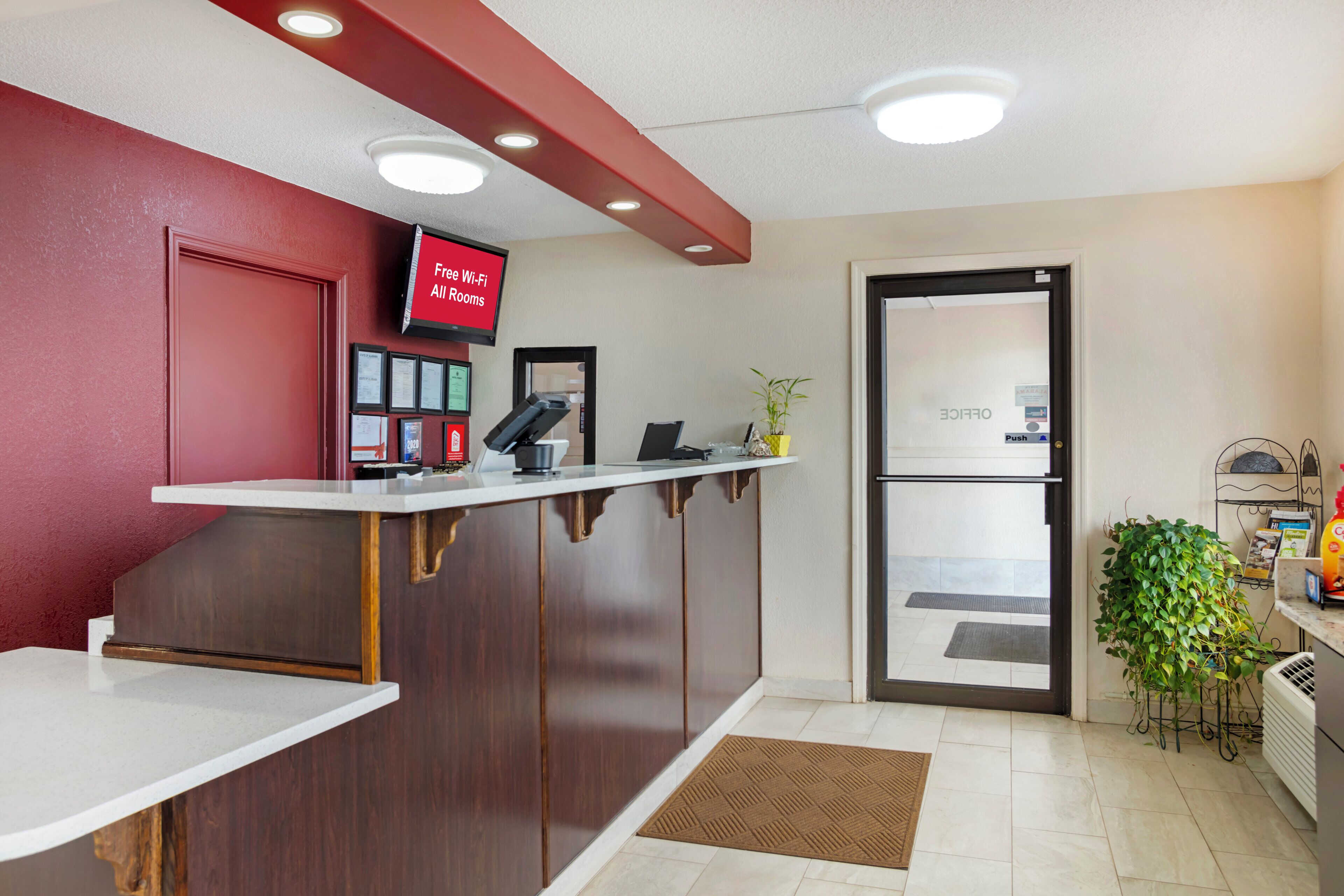Zona de recepción de un hotel, con mostrador de recepción, un televisor mostrando 'Wi-Fi gratuito en todas las habitaciones' y una puerta con la leyenda 'OFICINA'.