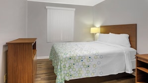 A hotel room with a bed, a nightstand, a wooden side table, a window with blinds, and a wall-mounted lamp. - HomeTowne Studios by Red Roof Huntsville - Madison (Madison)