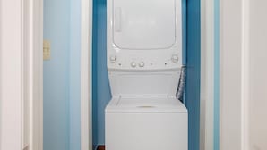 A compact, white washing machine in a narrow hallway with blue walls. - HomeTowne Studios by Red Roof Huntsville - Madison (Madison)