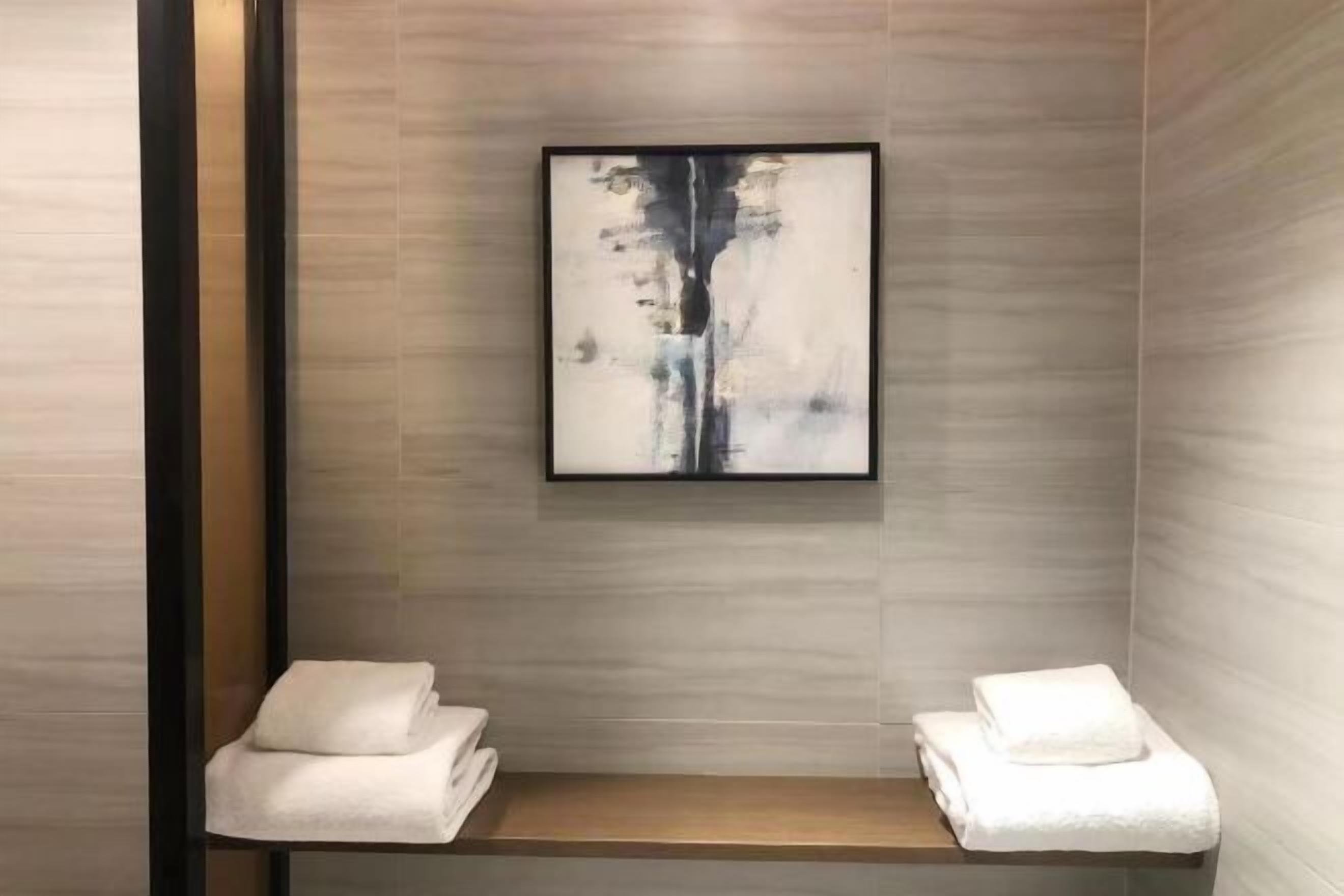 A modern bathroom with a minimalist design, featuring a wooden bench, two towels, and a framed abstract artwork on the wall.