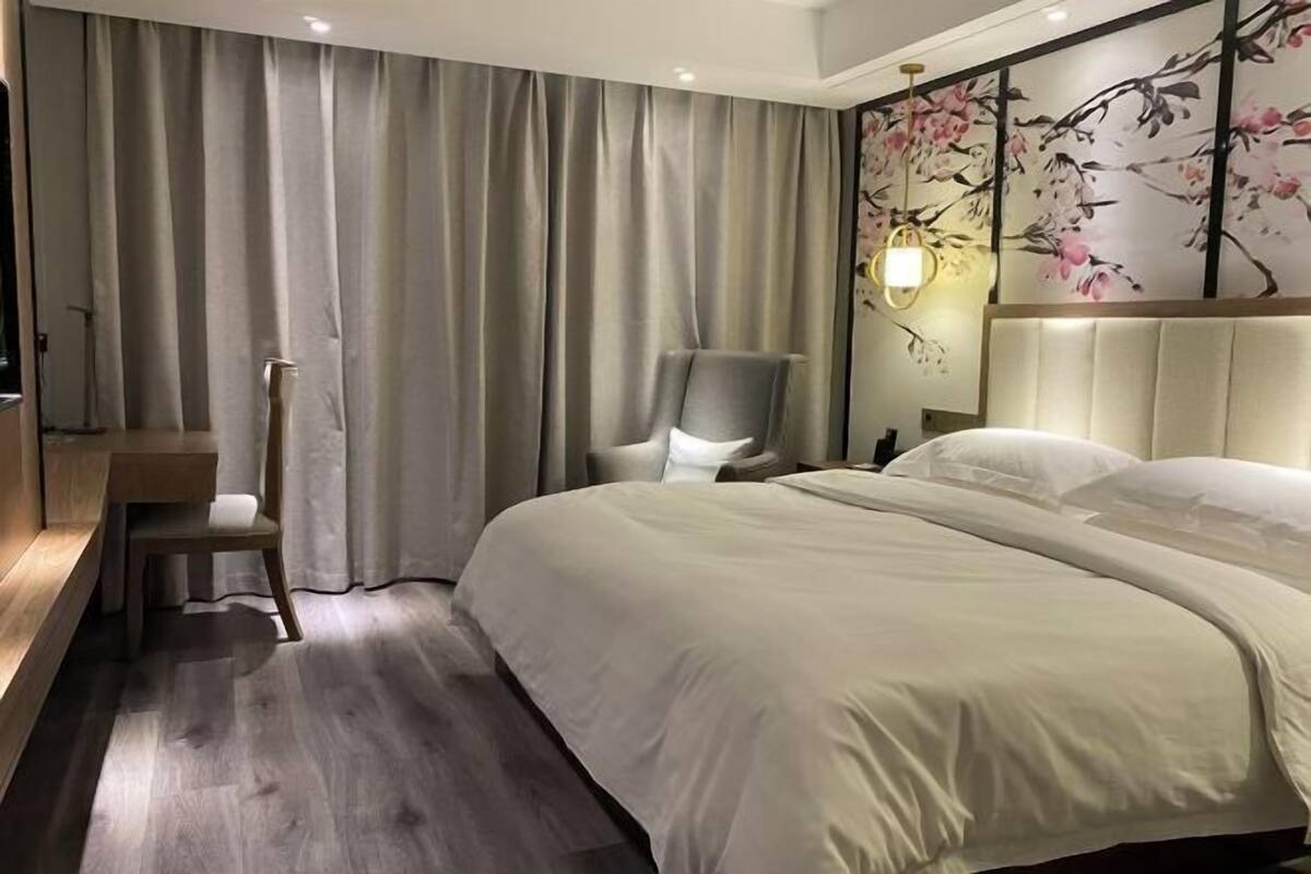 Quarto de hotel com uma cama grande, uma cadeira, uma escrivaninha e uma luminária fixada na parede.