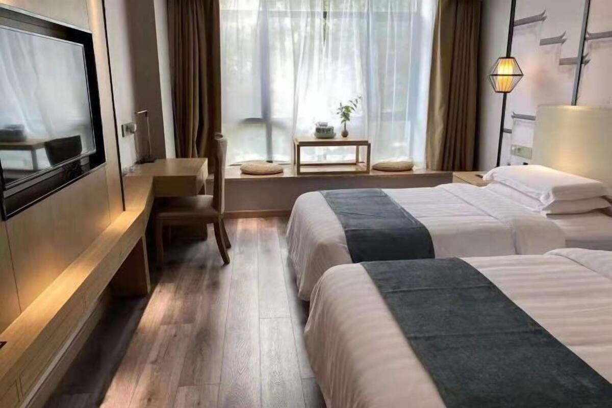 Quarto de hotel com televisão, duas camas, escrivaninha, cadeira, mesinha com planta e janela com cortinas.