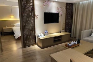 A modern hotel room with a wooden floor, a bed with white linens, a TV mounted on the wall, a wooden console table with a tray of cups, and a sofa.
