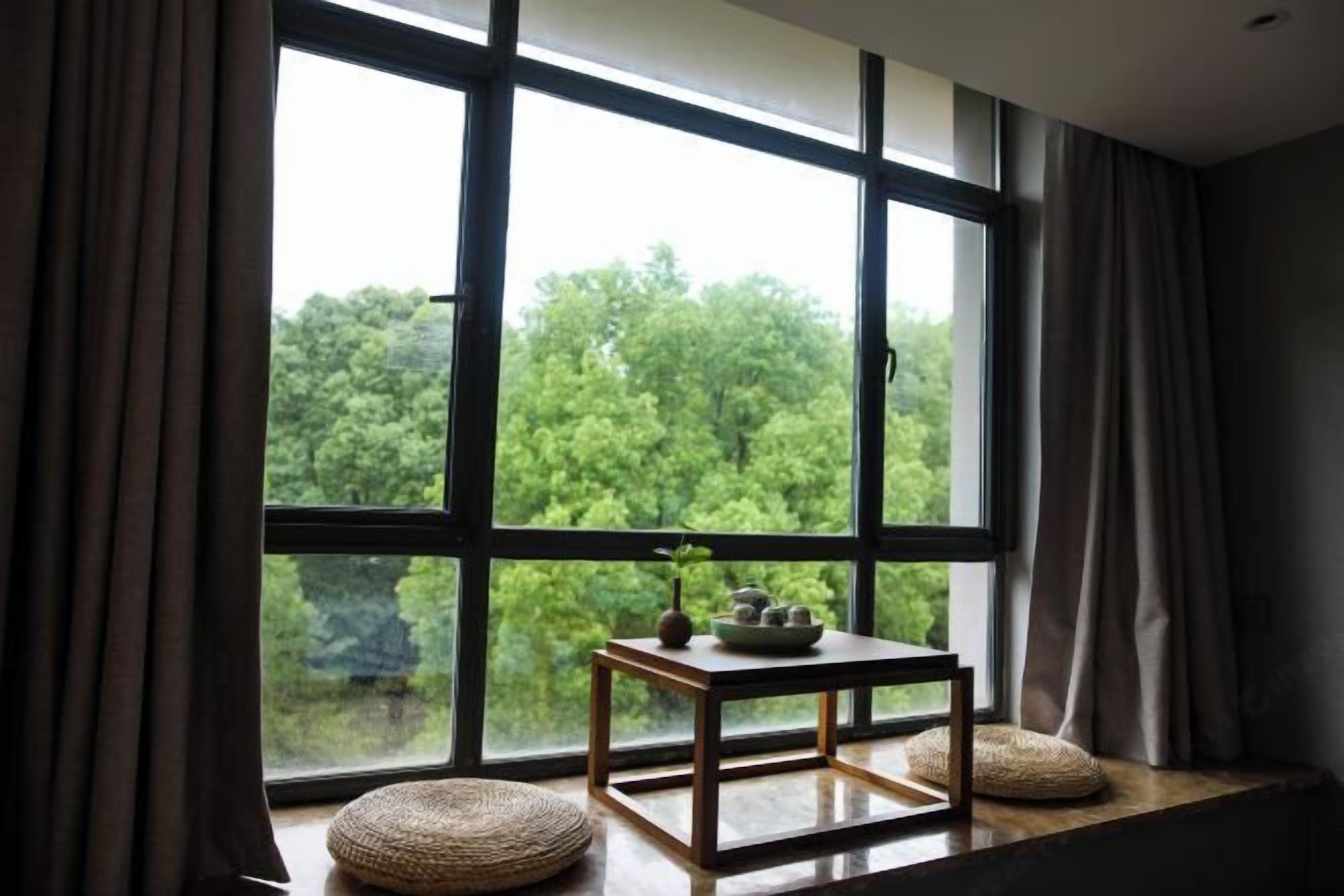 A room with a large window, a small table with a vase and teapot, and a woven cushion on the windowsill.