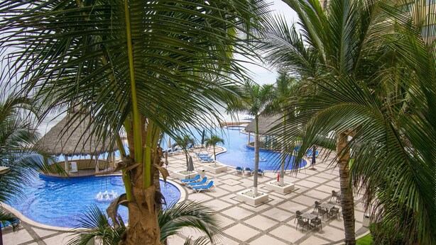 Un espace piscine dans un complexe hôtelier, comprenant plusieurs piscines, des chaises longues et des palmiers.