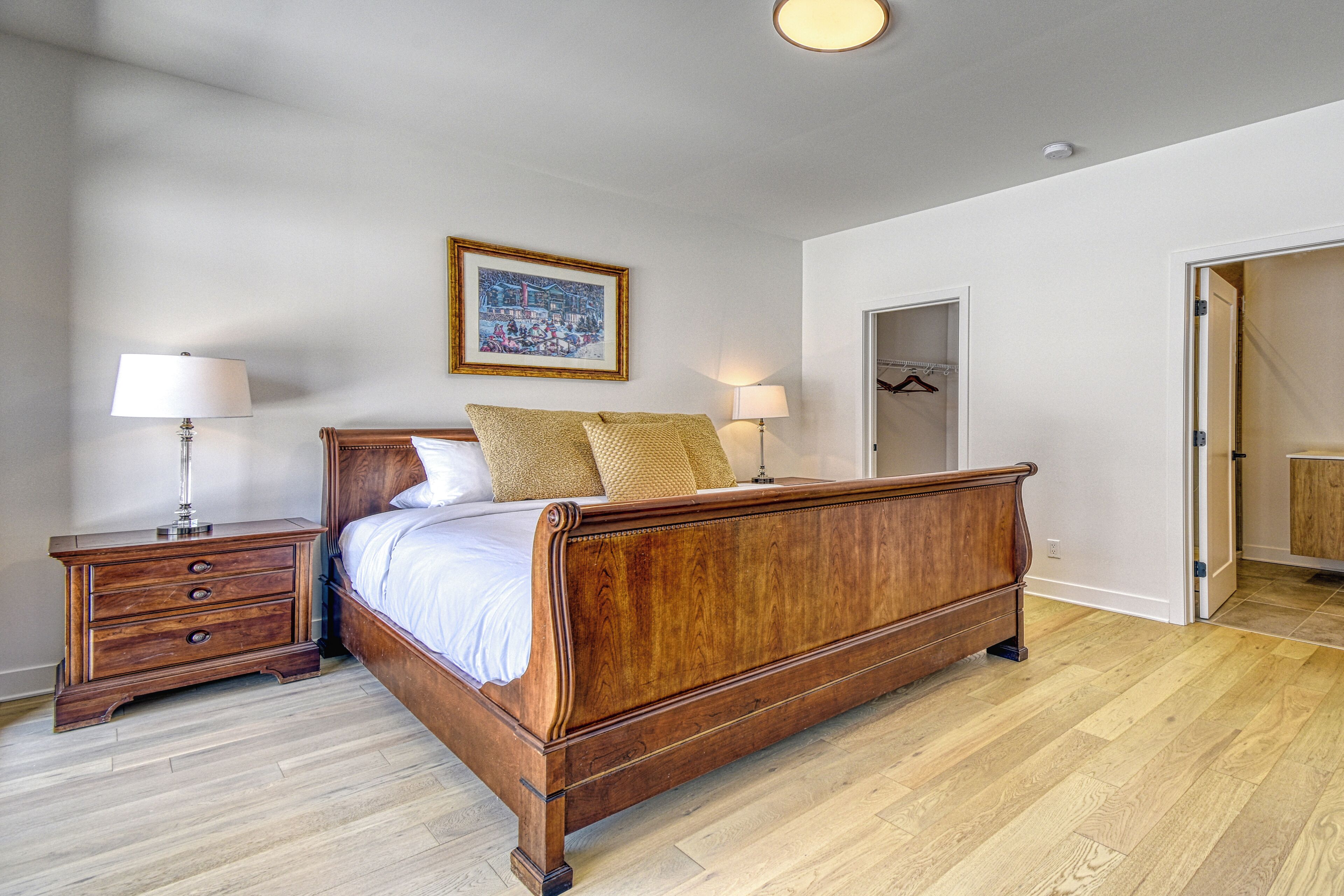 A bedroom with a wooden bed, two bedside tables with lamps, a framed picture on the wall, and a door leading to another room.