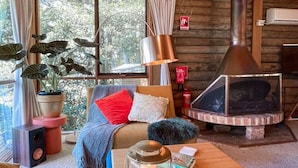 A cozy cabin interior with a wooden ceiling, a fireplace, a sofa with cushions, a coffee table, and a potted plant.