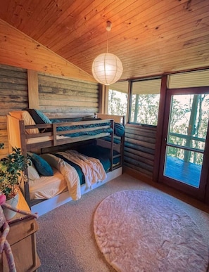 Een slaapkamer met een stapelbed, een houten plafond, een hanglamp, een raam met uitzicht op bomen, en een rond vloerkleed.