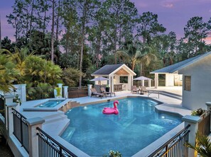 Un área de piscina con un flotador en forma de flamenco rosa, rodeada de palmeras y una casa con mobiliario exterior.