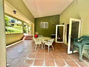 Varanda coberta com mesa redonda e cadeiras, uma porta e vista para um edifício.