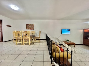 A spacious living room with a dining area, a sofa, a TV mounted on the wall, and a staircase leading to another level.