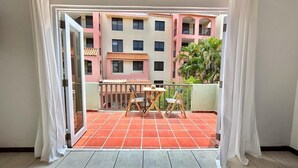 Uma varanda com piso de azulejos, mesa e cadeiras, com vista para um prédio com varandas e uma palmeira.