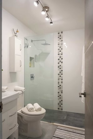 A modern bathroom with a glass shower enclosure, a white toilet, and a vanity with a mirror. - Beautiful Beach Studio @Condado (San Juan)