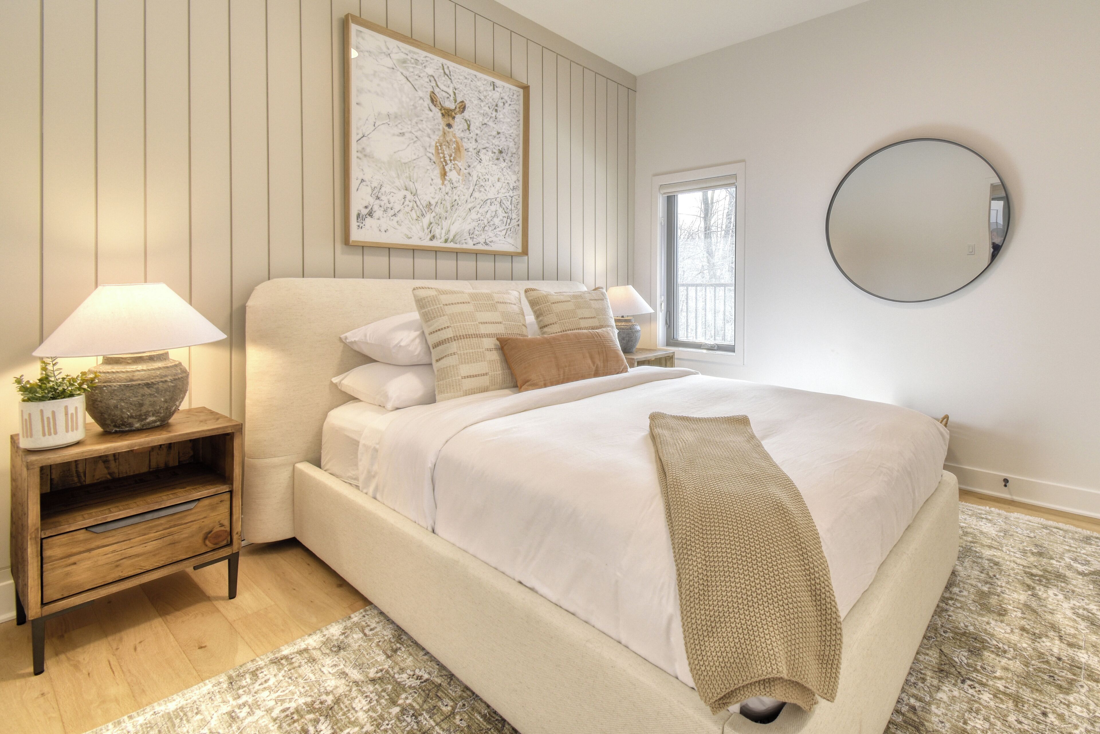 A bedroom with a large bed, a nightstand with a lamp, a framed picture on the wall, and a round mirror.