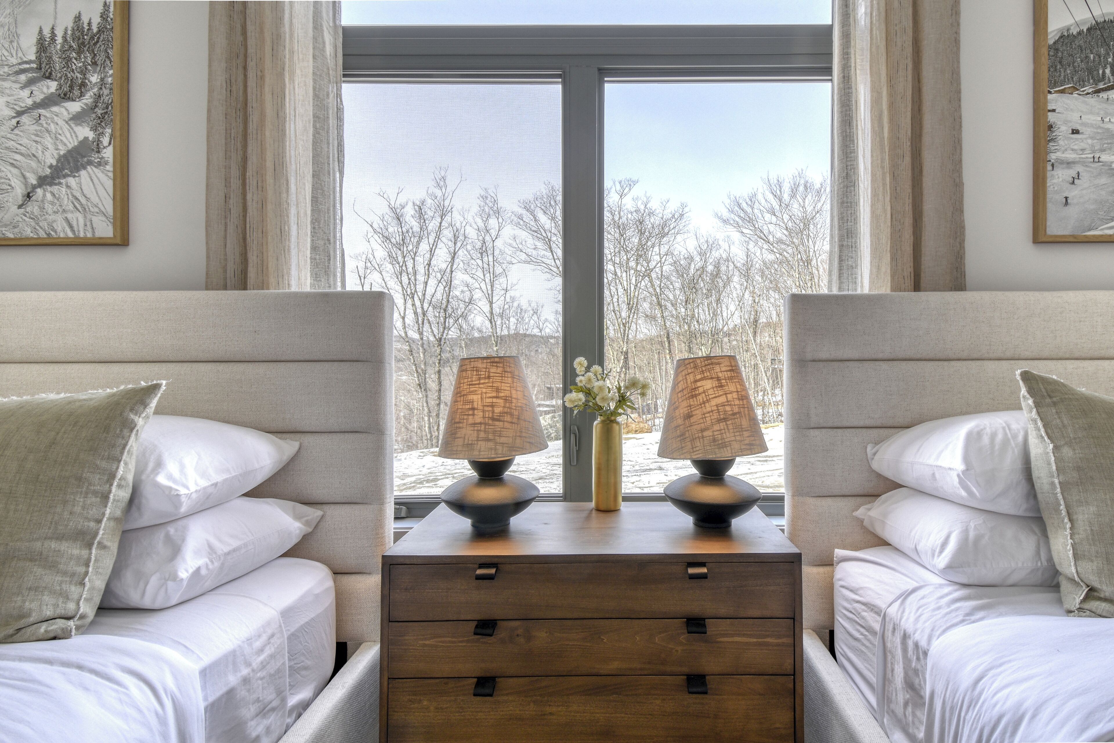 A hotel room with two beds, a wooden nightstand with lamps, and a large window with a view of trees and hills.