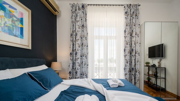 A hotel room with a bed, a TV, a shelf with items, and a painting on the wall.