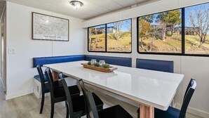 Un espace repas avec une table blanche, des bancs bleus et de grandes fenêtres donnant sur un parc.