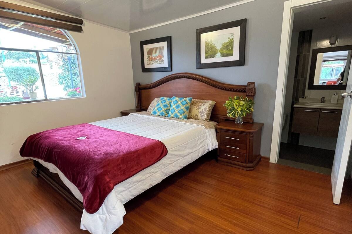 Un dormitorio con cama de madera, una ventana y un baño visible a través de una puerta abierta.