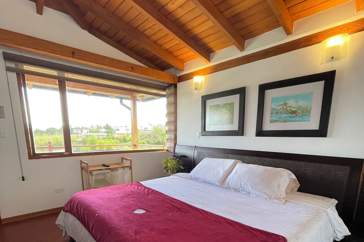 Un dormitorio con una cama grande, vigas de madera en el techo, una ventana con vistas a la vegetación y dos cuadros en la pared.