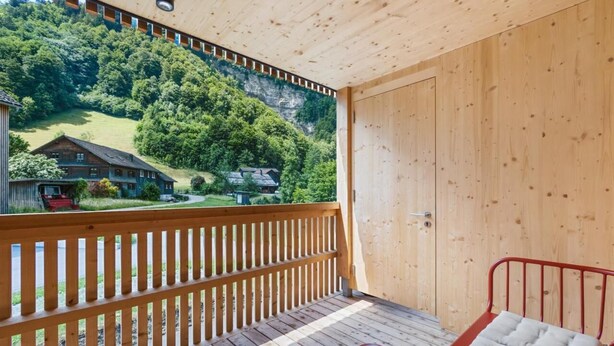 A wooden balcony with a view of a house and forested hillside. - Appartement Johann - Mellau - Urlaub im Herzen des Bregenzerwaldes (Mellau)