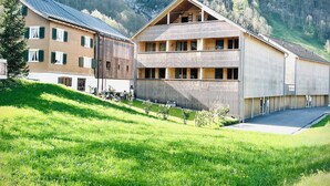 A two-story building with a balcony, surrounded by a grassy area and a forested hillside. - Appartement Johann - Mellau - Urlaub im Herzen des Bregenzerwaldes (Mellau)