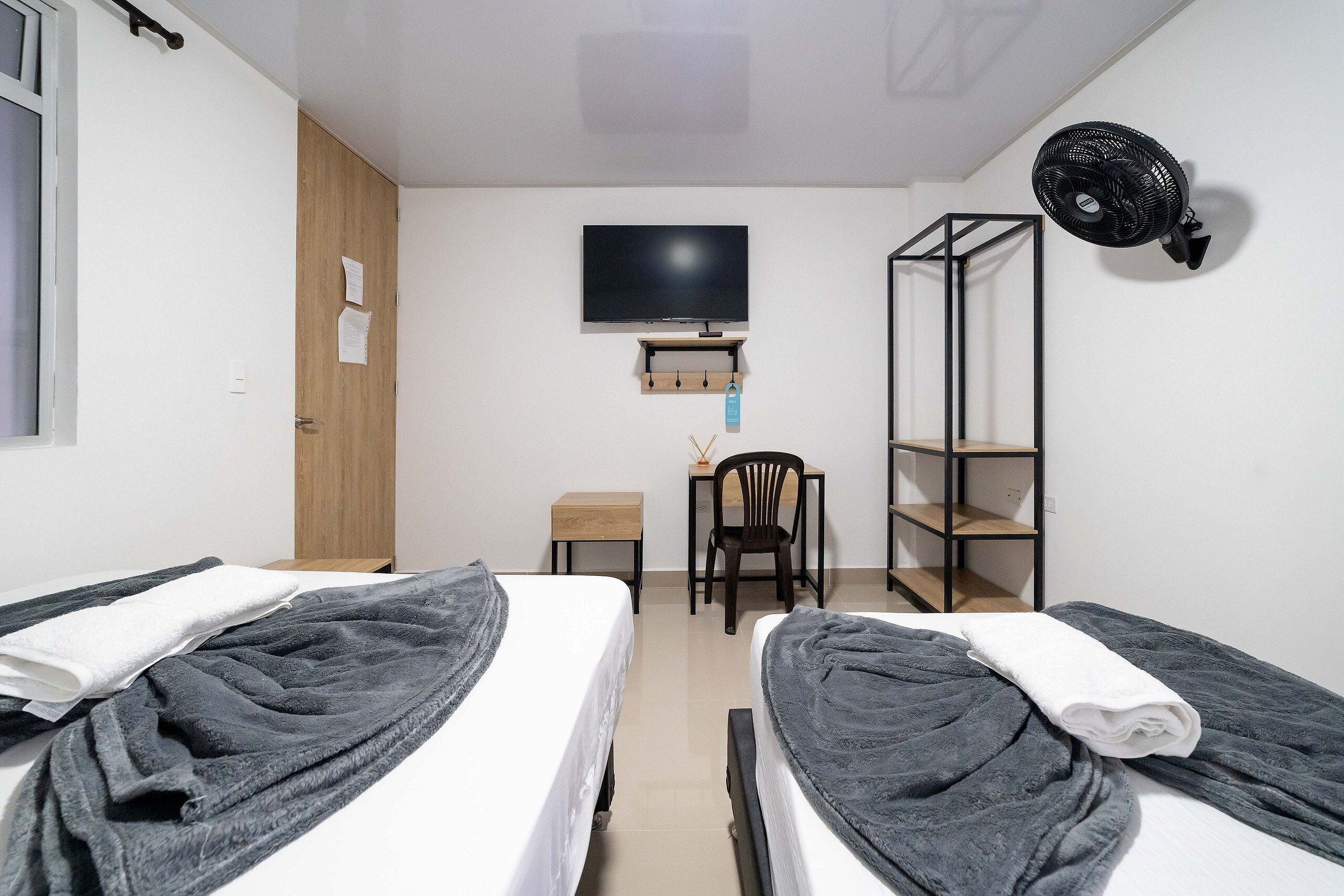 A room with two beds, a TV, a fan, a chair, and a small table.