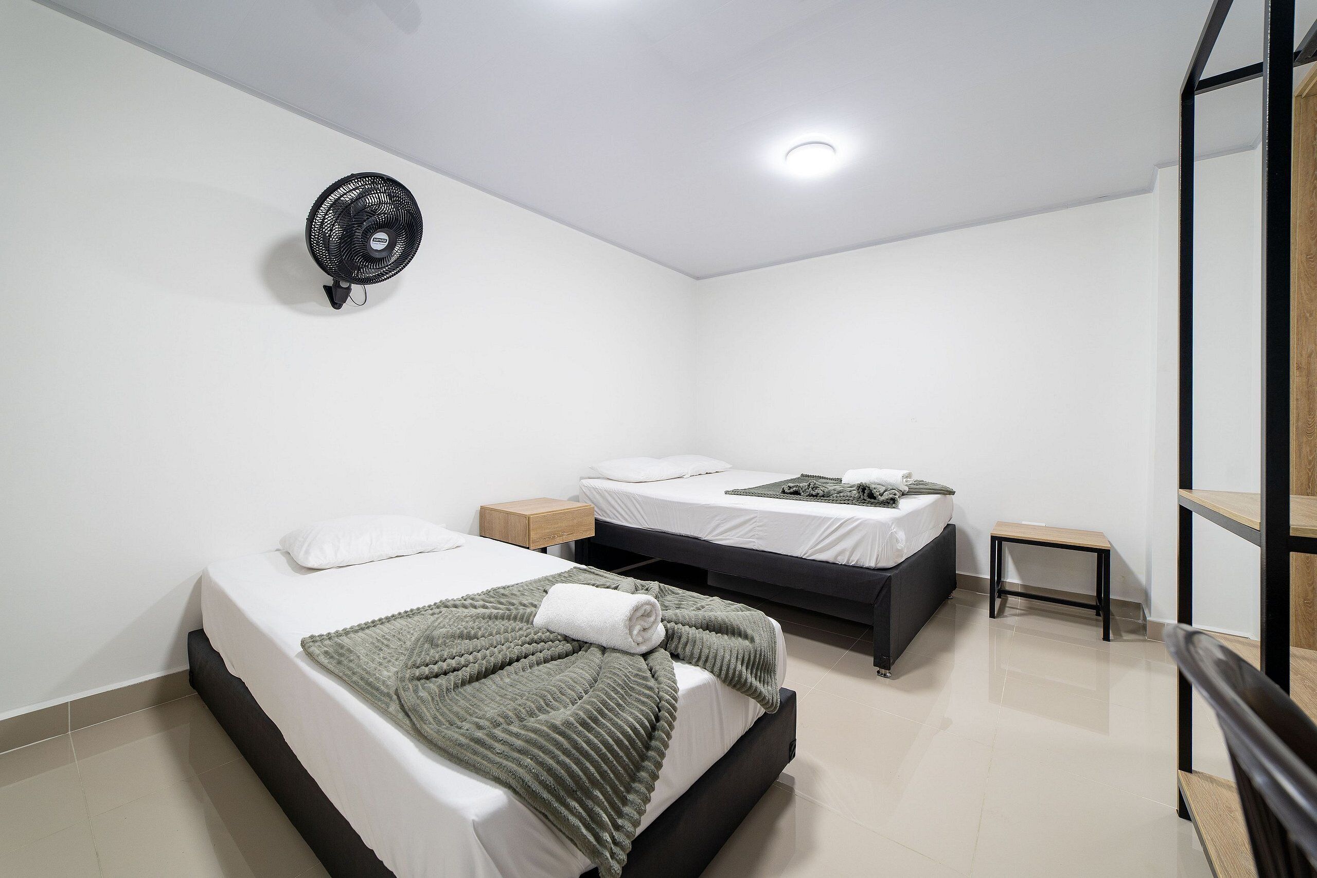 Una habitación de hotel moderna con dos camas, una mesita de noche pequeña, un ventilador de pared y un banco de madera.