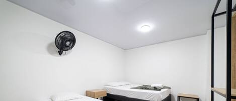 Una habitación de hotel moderna con dos camas, una mesita de noche pequeña, un ventilador de pared y un banco de madera.