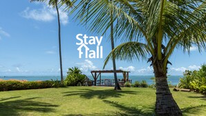 A tropical garden with a palm tree, a wooden pergola, and a view of the ocean. - Beira Mar-Cozinheira-Piscina-4QT-SH029 (Sirinhaem)