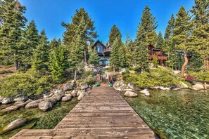 Eine Holzterrasse führt zu einer Lodge, umgeben von hohen Bäumen und einem klaren Gewässer.