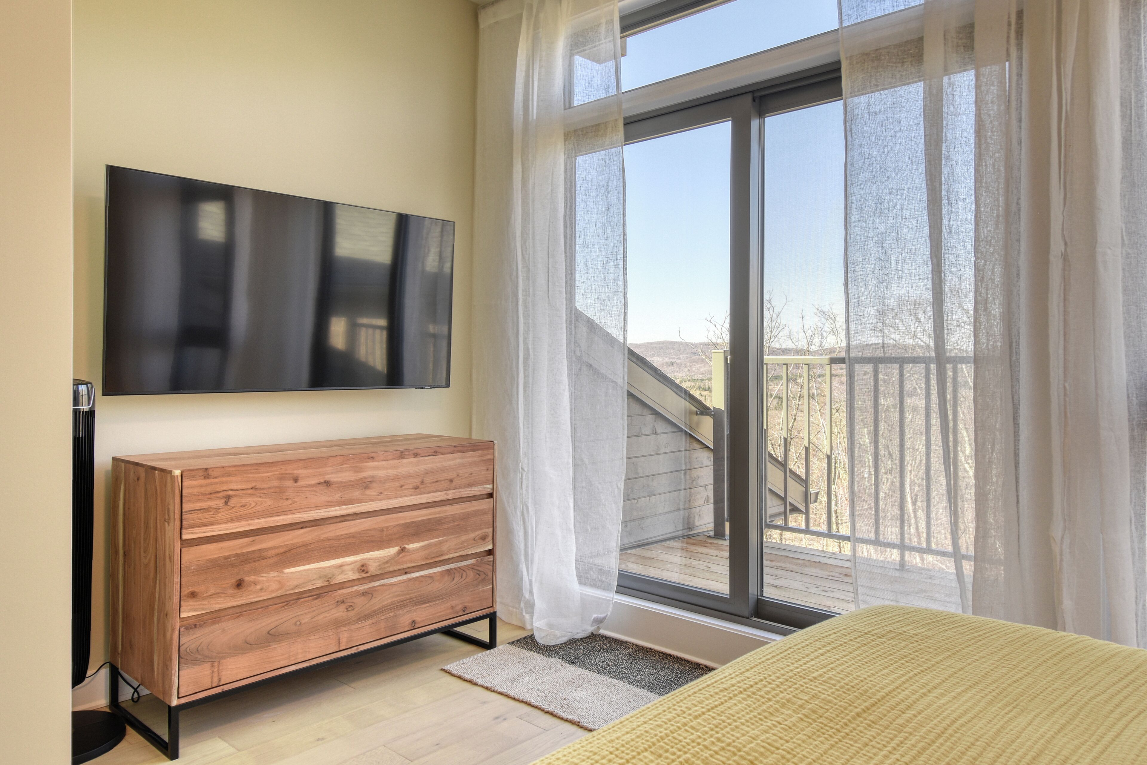 Quarto com cômoda de madeira, TV de tela plana, janela grande com cortinas leves e vista para uma varanda.