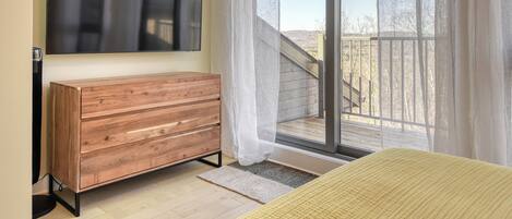 Een slaapkamer met een houten dressoir, een flatscreen televisie, een groot raam met dunne gordijnen en uitzicht op een balkon.