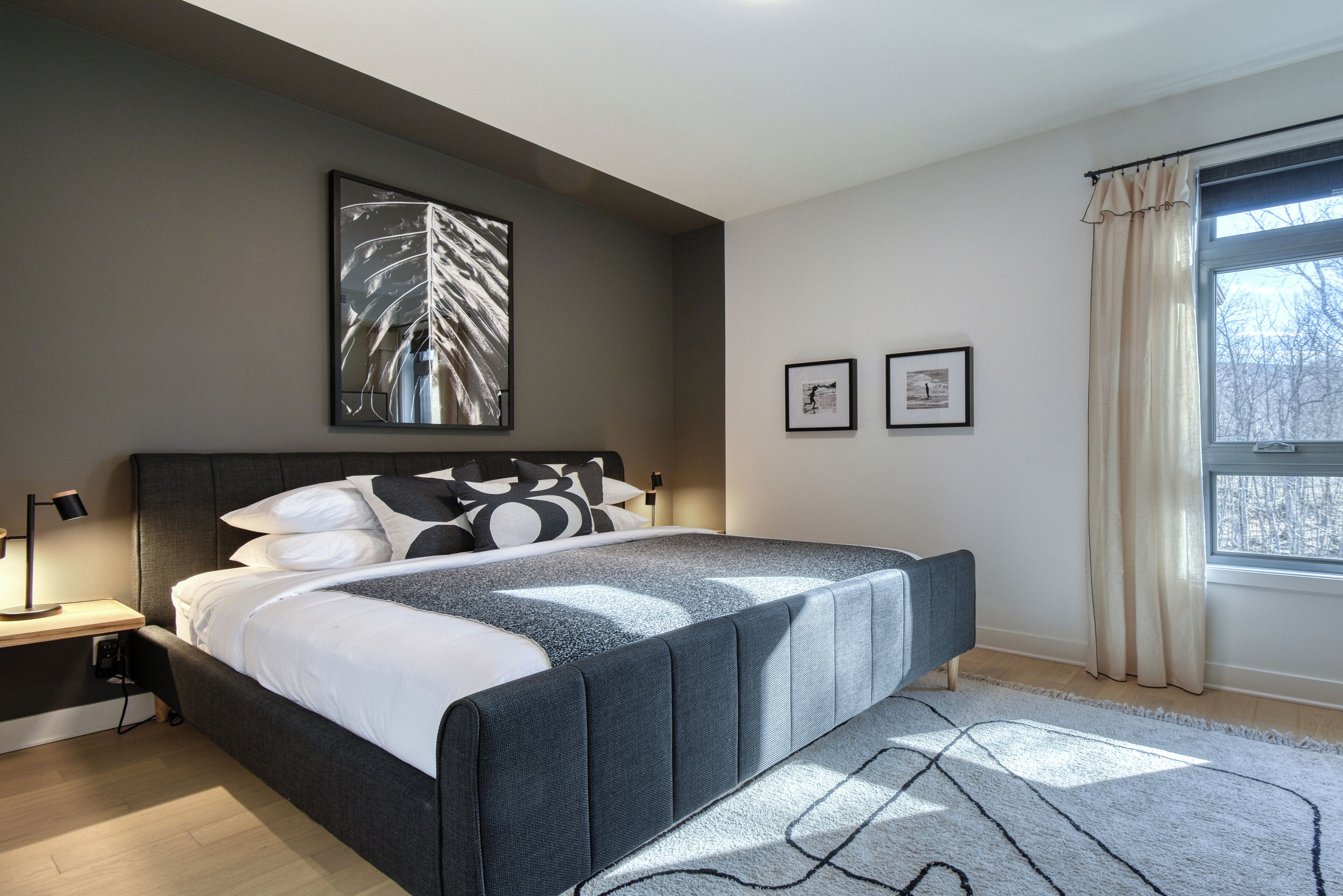 A modern bedroom with a large bed, a nightstand, a wall-mounted lamp, and a window with curtains.