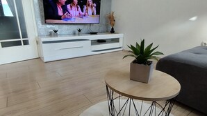 A modern living room with a flat-screen TV on a white entertainment unit, a round coffee table with a plant, and a grey sofa.