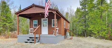 A wooden cabin with a porch, an American flag, and a dirt path.