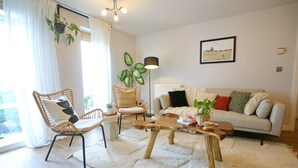 Ein Wohnzimmer mit einer Couch, zwei Sesseln, einem Couchtisch und einem großen Fenster mit Vorhängen.