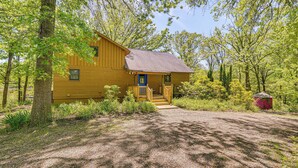 A yellow cabin with a porch and steps, surrounded by trees and a gravel path. - Historic Clark Cabin w/ Deck & Fishing Pond Access (Clark)
