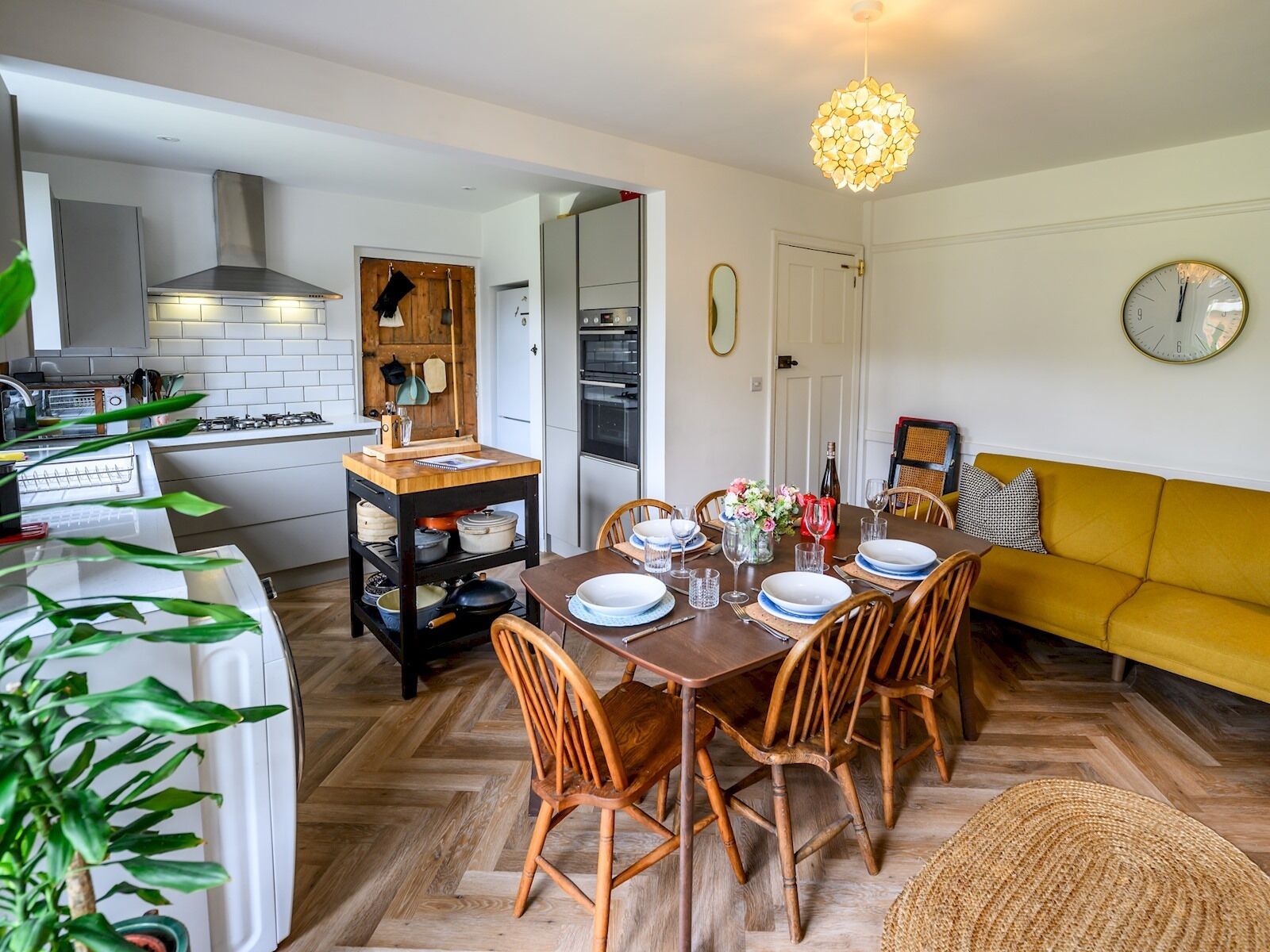 Een moderne keuken met witte kasten, een houten eettafel voor vier personen en een gele bank in de woonkamer.