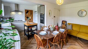 Een moderne keuken met witte kasten, een houten eettafel voor vier personen en een gele bank in de woonkamer.