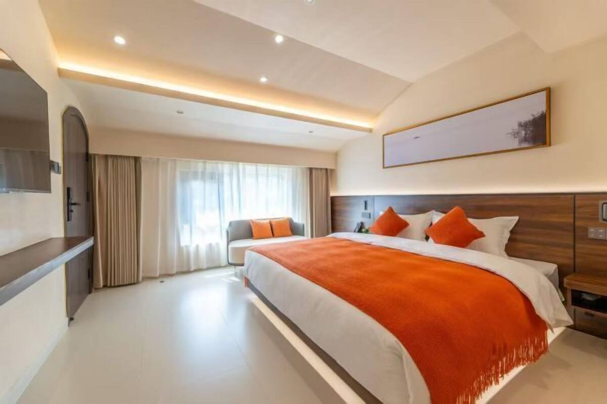 A modern hotel room with a large bed, a flat-screen TV, a sofa, and a large window with curtains.